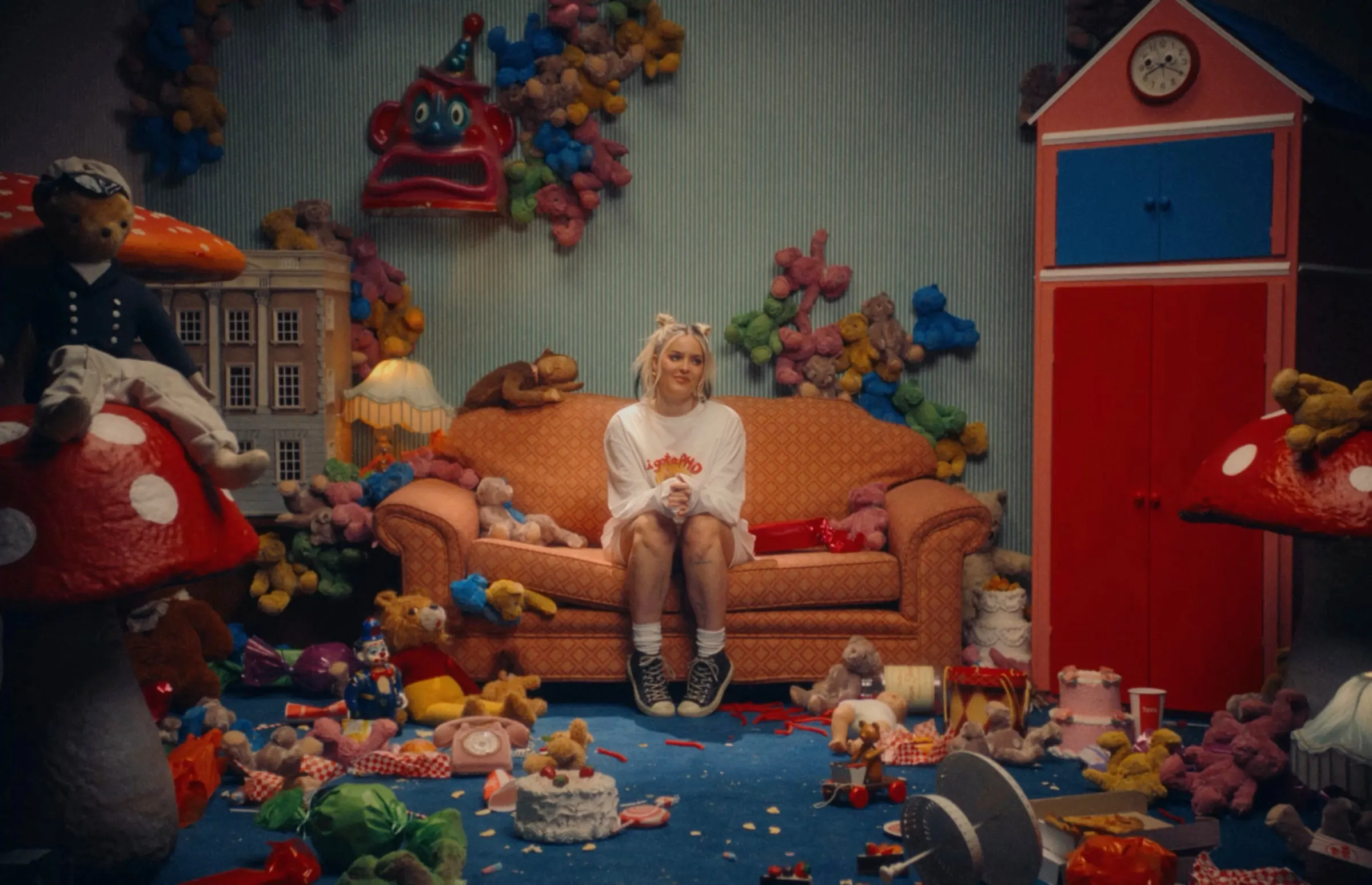 Anne-Marie on an orange sofa in a toy-strewn, candy-coloured living room, from the Unhealthy album visual
